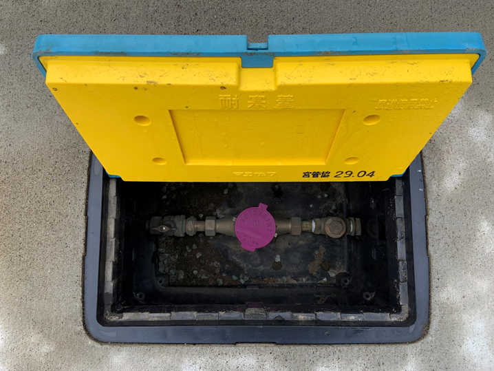 地面に設置されたメーターボックスの水色の蓋を開け、蓋の内側についた黄色の保温材と、地中に収められた水道メーターが見える写真