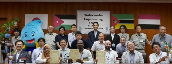 日本、ヨルダン、モザンビーク、イエメンの国旗が掲げられた壁を背景に、修了証を手にした派遣研究員の方々が、ウォッターくん、関係者と一緒に並んだ閉校式の記念写真