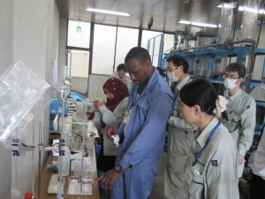 ビーカーや検査器具が並ぶ台の前で、派遣研究員が職員の説明を受けながら薬品注入率の計算を行っている様子の写真