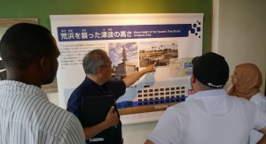 「荒浜を襲った津波の高さ」と記されたパネルに集まった派遣研究員へ、男性が説明を行っている震災遺構荒浜小学校見学の様子の写真