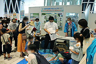 たくさんの親子連れでにぎわうイベント会場で、複数のポスターなどが貼られた白いパネル前に設けられたスーパーボールすくいの小さなビニールプールの周りに子どもたちが集まっている様子の写真