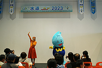 「水道フェア2023」の横断幕が掲げられた壁の前で、オレンジ色の衣装を着た女性と水滴をモチーフにしたキャラクターのウォッターくんが、客席に座った観客に向けてポーズをとっている写真