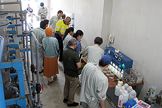 配管設備のある屋内の作業場で、派遣研修員の人々が薬品注入率の計算机に並べられた水質検査用のビーカーや機材を熱心に見学している写真