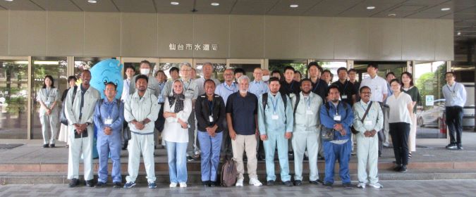 仙台市水道局の建物の前で、様々な国籍のJICA派遣研修員の男女のグループがウォッターくんのマスコットと一緒に並んで笑顔で立っている到着時の集合写真
