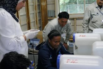 複数並んだ白いポリタンクの前で、派遣研究員の方々が職員の説明を受けながら薬品手動注入に取り組んでいる様子の写真