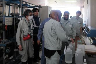 複数のポリボトルや青い実験装置が並ぶ台の前で、派遣研究員が薬品注入率の計算について話し合っている様子の写真