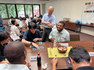 テーブルが並んだ明るい室内で、お菓子や飲み物を前に着席した派遣研究員の方々が、職員たちと交流している様子の写真