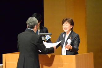 式典の壇上で、笑顔を浮かべた郡市長から、黒いスーツ姿の男性へ表彰状の授与が行われている様子の写真