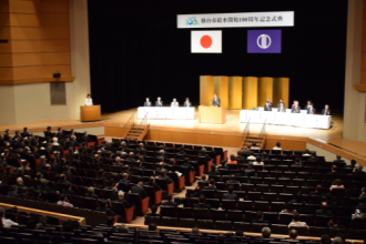 日の丸、市旗が掲げられた広いホールの壇上に、金屏風を背にして中央に演台、左右に来賓席が設けられ、多くの出席者が観客席に着席している式典会場全体の様子の写真
