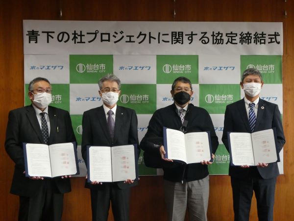 「青下の杜プロジェクトに関する協定締結式」と書かれ、仙台市と「水のマエザワ」のロゴが交互に並ぶパネルの前で、マスクを着用した4名の男性が締結した協定書を手に持ち並んでいる記念写真