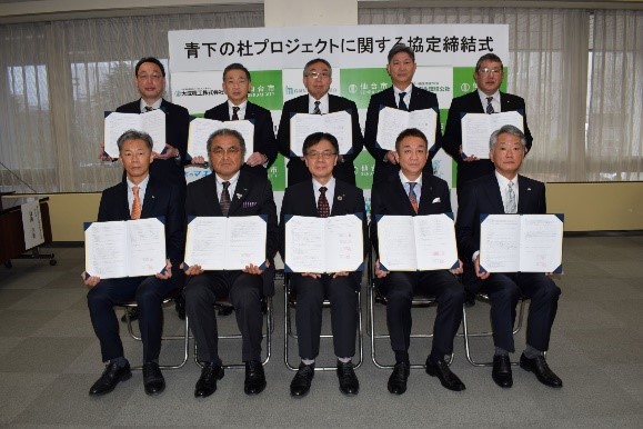 「青下の杜プロジェクトに関する協定締結式」と書かれたパネルの前で、スーツ姿の男性10名が締結した協定書を広げて持ち、前後2列に並んだ記念写真