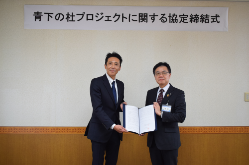 「青下の杜プロジェクトに関する協定締結式」と書かれた横断幕の前で、スーツ姿の男性二人が笑顔で並び、協定書を正面に向けて一緒に持っている記念写真