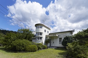 円筒形の監視塔のような造りや丸窓が特徴的な白い洋風建築物が、手入れされた芝生や青々とした植栽に囲まれて建つ、モダンな雰囲気を感じさせる旧管理事務所の写真