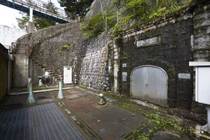 苔むした重厚な石積みのトンネルの入り口に金属製の頑丈な扉が設置され、傍らには古い配管設備や点検用の階段、その上部に架かる管理橋が見える青下隧道入口の写真