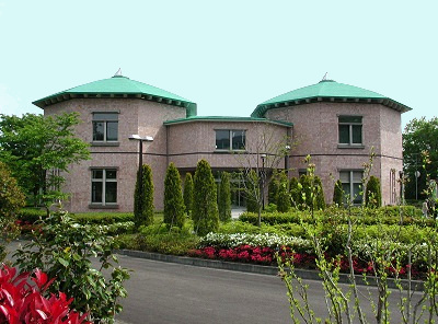 手前に色とりどりの花壇や円錐形に整えられた植栽が並び、緑色の屋根とレンガ調の外壁を持つ多角形の建物が連結した水道記念館の外観写真