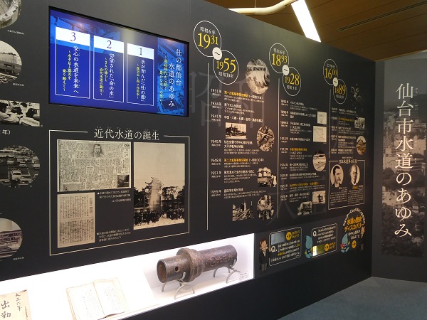 「仙台市水道のあゆみ」と題された壁面展示に、1600年代から1955年までの歴史年表や「近代水道の誕生」の解説パネル、モニター映像、そして古い水道管の実物が展示されている写真