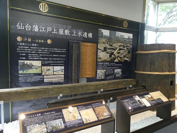 「仙台藩江戸上屋敷 上水遺構」と記された大型の解説パネルの前に、発掘された木製の水道管や大きな木桶が展示されている写真