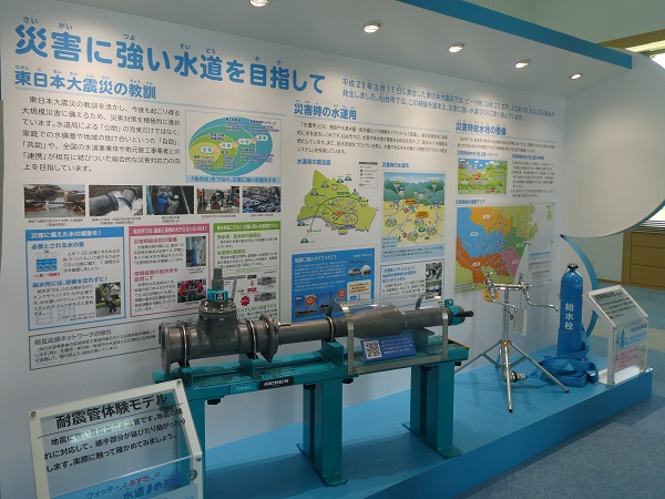 「災害に強い水道を目指して」という見出しのもと、東日本大震災の教訓や災害時の水運用を解説するパネルとともに、地震の揺れを吸収する「耐震管体験モデル」や青い給水栓などの資機材が展示された写真