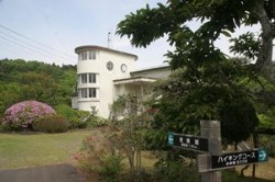 円筒形の塔のような外観を持つ白い建物の手前に「ハイキングコース」などの案内看板やピンク色の花が咲く植栽が広がる写真