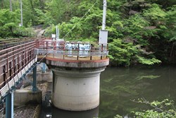 緑豊かな山林を背景に、水面にせり出した円柱状のコンクリート構造物と、それに続く手すり付きの管理用通路が設置されたダム施設の写真