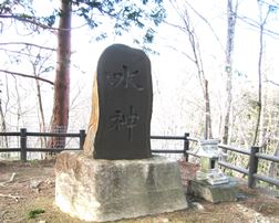「水神」と刻まれた大きな黒い石碑が台座の上に立ち、その傍らに小さな石造りの祠が置かれた写真