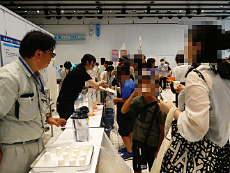 屋内のイベント会場にて、作業服を着たスタッフが並べられた紙コップやピッチャーを前に、来場した親子連れなどに説明や案内を行っている様子の写真