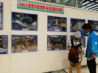「令和6年 能登半島地震 応援活動写真展」のパネル展示にて、法被を着たスタッフが支援活動の様子を写した写真を指差しながら来場者の子供に熱心に説明している様子の写真
