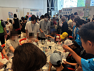 賑やかな屋内イベント会場にて、多くの子供たちがテーブルを囲み、スタッフのサポートを受けながらペットボトルやボウルを使って実験や工作に熱心に取り組んでいる写真