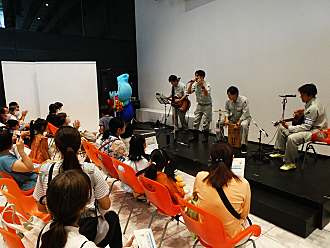 屋内のステージにて、作業服を着たバンドメンバーがギターやカホンを演奏しながら歌い、着ぐるみのウォッターくんが見守る中で、椅子に座った親子連れなどの観客が手拍子をして楽しんでいる写真