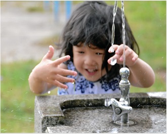 公園の水飲み場から勢いよく噴き出す水に、笑顔の子供が楽しそうに手を伸ばして触れようとしている写真