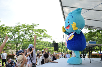 屋外のステージ上で、ウォッターくんが笑顔で手を振り、それを見上げる多くの子供たちが元気よく手を挙げて応えている、イベント会場での写真