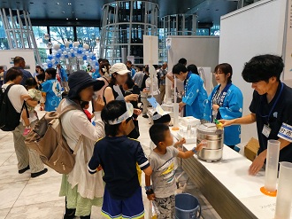 青い法被を着たスタッフたちが、ブースに集まった親子連れにコップを手渡し、ウォータージャグから注いだ水を試飲してもらっている写真