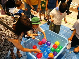 水に浮かんだカラフルなヨーヨーを、子どもたちが釣り針のついた紐を使って釣り上げようと楽しんでいる写真