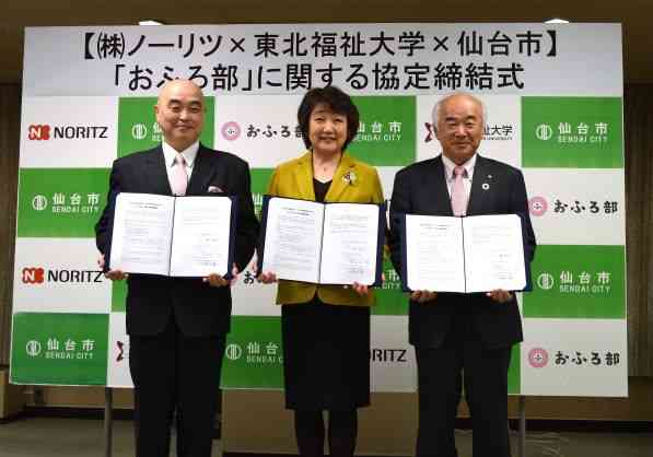「株式会社ノーリツ&times;東北福祉大学&times;仙台市」による「おふろ部」に関する協定締結式にて、各組織のロゴが並ぶ背景の前で、スーツ姿の代表者3名が署名済みの協定書を手に持ち記念撮影している写真
