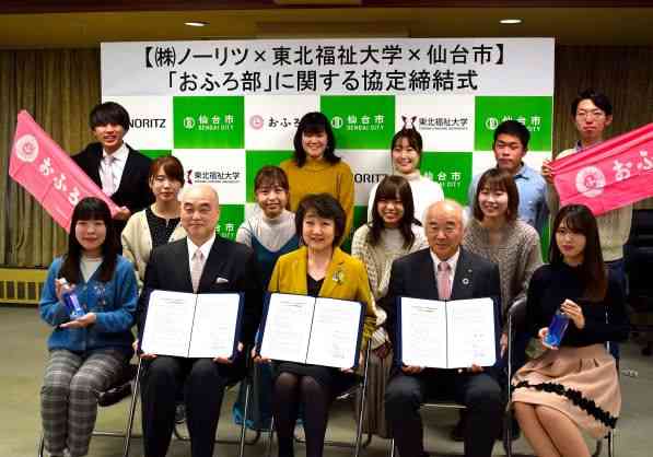 「株式会社ノーリツ&times;東北福祉大学&times;仙台市」の「おふろ部」に関する協定締結式にて、署名した協定書を持つ代表者3名を中心に、ピンク色のタオルを掲げる学生たちを含む総勢14名が、各組織のロゴが並ぶ背景の前で笑顔で記念撮影をしている写真
