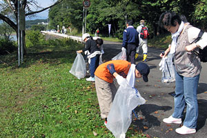 道路脇で、軍手をはめて大きな透明のゴミ袋を手にした子どもたちや大人たちが、落ちているゴミを拾い集めながら清掃活動に励んでいる様子の写真