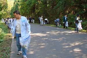 木々が立ち並ぶ舗装路で、多くの人々がゴミ袋を手に持ちながら左右に分かれて路肩のゴミを拾い集めている様子の写真