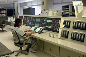 多数のモニターや計器が並ぶ大規模な制御盤の前に作業員が座り、施設の稼働状況を監視している管理室の写真