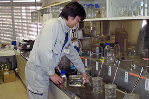 室内で、作業着を着た男性が水道の5つ蛇口から流れる水をビーカーなどの容器に入れ、プラスチックのボトルの水を捨てている様子の水質検査の写真