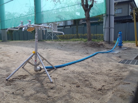 砂地に設置された青い給水栓から青いホースが伸び、三脚で自立する複数の蛇口を備えた仮設給水装置に接続されている様子の写真