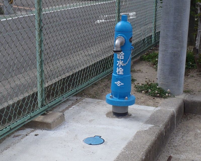 「給水柱」の文字が縦書きで記された金属製ノズル付きの青い筒状の給水柱を写した写真