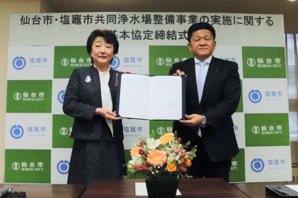 「仙台市・塩竈市共同浄水場整備事業の実施に関する基本協定締結式」において、両市のロゴが並ぶバックパネルの前で、仙台市長と塩竈市長が署名済みの協定書を手に持ち、並んで立つ記念写真