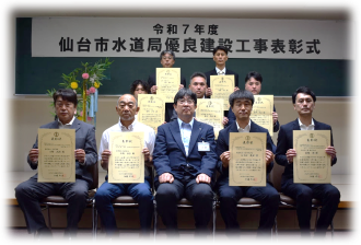 「令和7年度 仙台市水道局優良建設工事表彰式」と書かれた横断幕を背に、賞状を手にしたスーツ姿の方9名が、前列中央に座ったスーツ姿の男性と一緒に、前後3列に並んだ記念写真