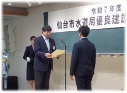 スーツ姿の男性がマイクに向かって手に持った表彰状を読み上げ、対面に受賞者の男性が立っている表彰状授与の様子の写真