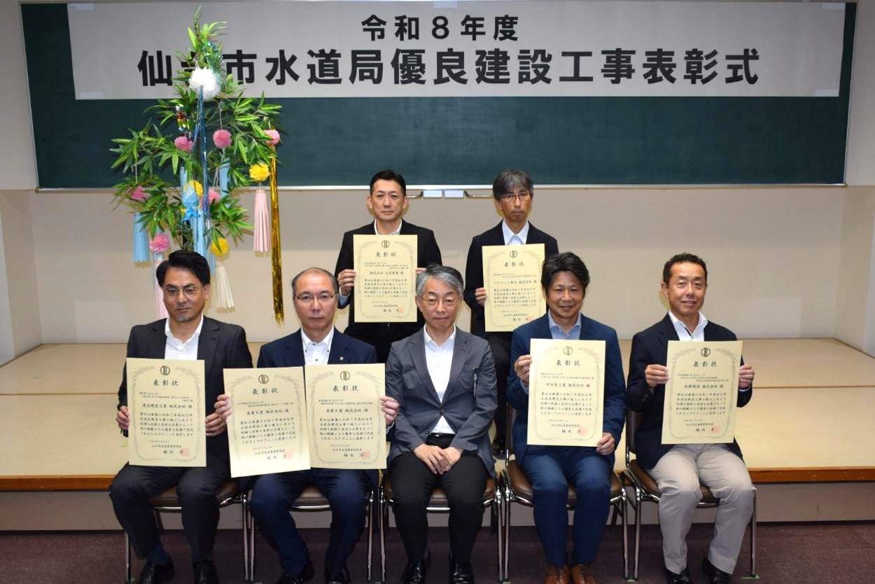 「令和8年度 仙台市水道局優良建設工事表彰式」と書かれた横断幕を背に、賞状を手にしたスーツ姿の方6名が、前列中央に座ったスーツ姿の男性と一緒に、前後2列に並んだ記念写真