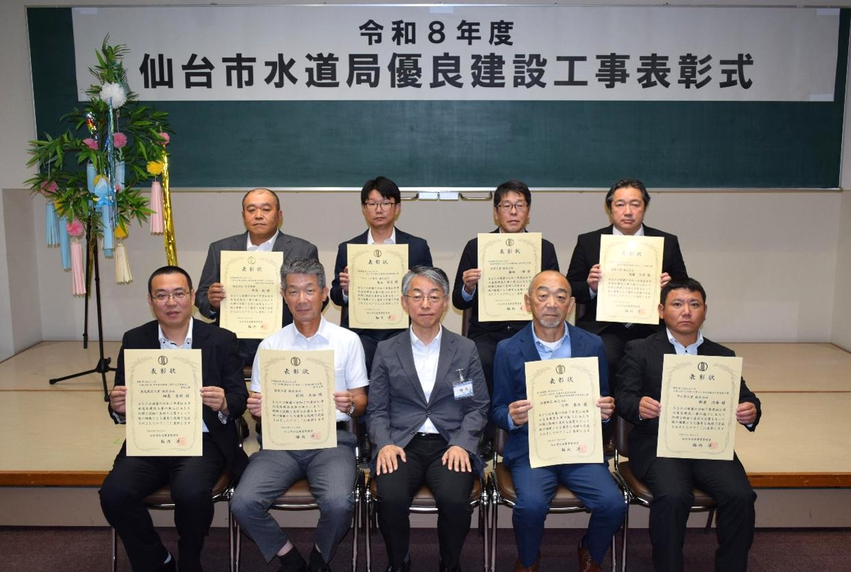 「令和8年度 仙台市水道局優良建設工事表彰式」と書かれた横断幕を背に、賞状を手にしたスーツ姿の受賞者8名が、前列中央に座ったスーツ姿の男性と一緒に、前後2列に並んだ記念写真