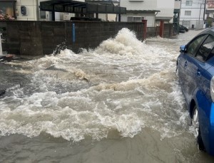 住宅の塀付近の地面から激しく水が噴き出し、冠水した道路には青い乗用車のタイヤが浸かるほどの濁った水が押し寄せている様子の写真